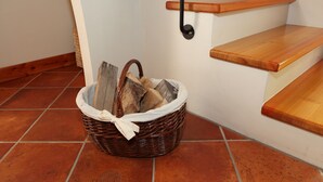 Uma cesta de vime com lenha sobre um chão de azulejos, ao lado de uma escada com degraus de madeira e prateleiras pretas fixadas na parede.