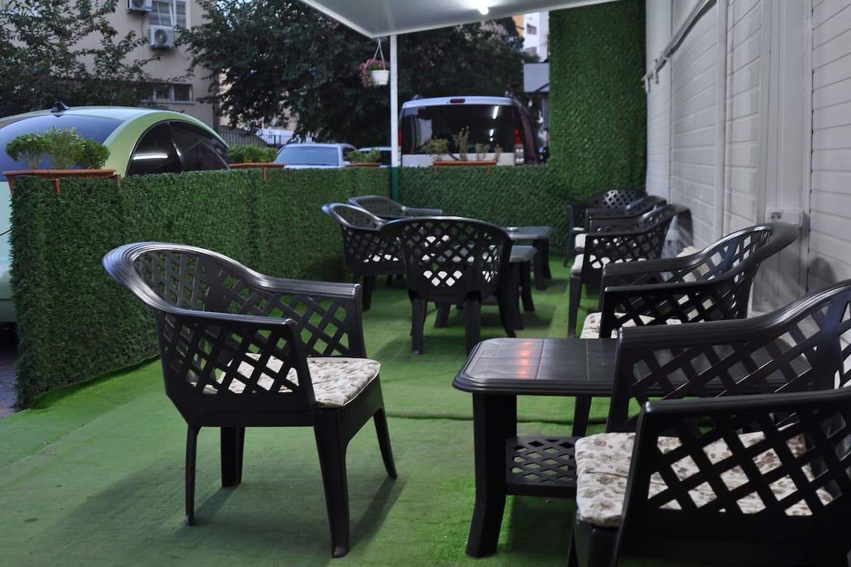 Un área de estar al aire libre con sillas y mesas negras, rodeada por un seto verde y una pared blanca.
