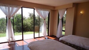 A room with two beds, large windows with white curtains, and a view of greenery outside. - Marula House (Naivasha)