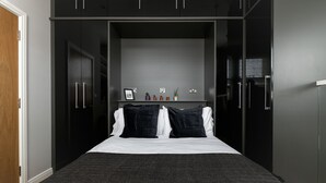 A compact bedroom with a bed, dark wardrobe, and a small shelf with decorative items. - 3 Beds 2 Baths Penthouse (London)