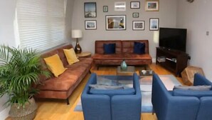 A living room with a brown sofa, blue armchairs, a coffee table, and a TV. There are framed pictures on the wall and a potted plant. - 3 Beds 2 Baths Penthouse (London)