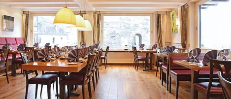 Un restaurante con mesas y sillas de madera, ventanas amplias y lámparas colgantes.