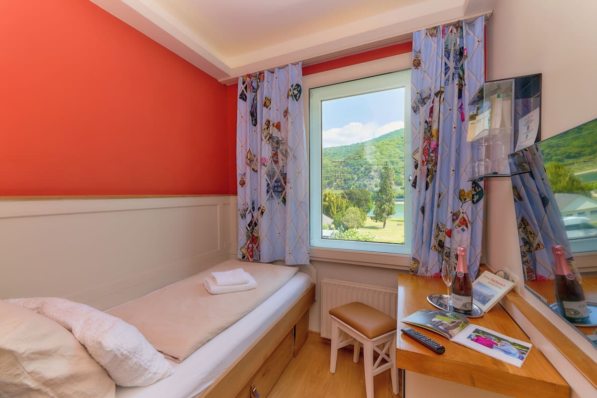 A single bed with a white blanket, a small wooden stool, a desk with a bottle, a remote control, and a framed picture.