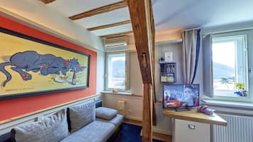 Een gezellige kamer met een houten plafond, een grijze bank, een schilderij van een kat, een bureau met een computer en een raam met uitzicht.