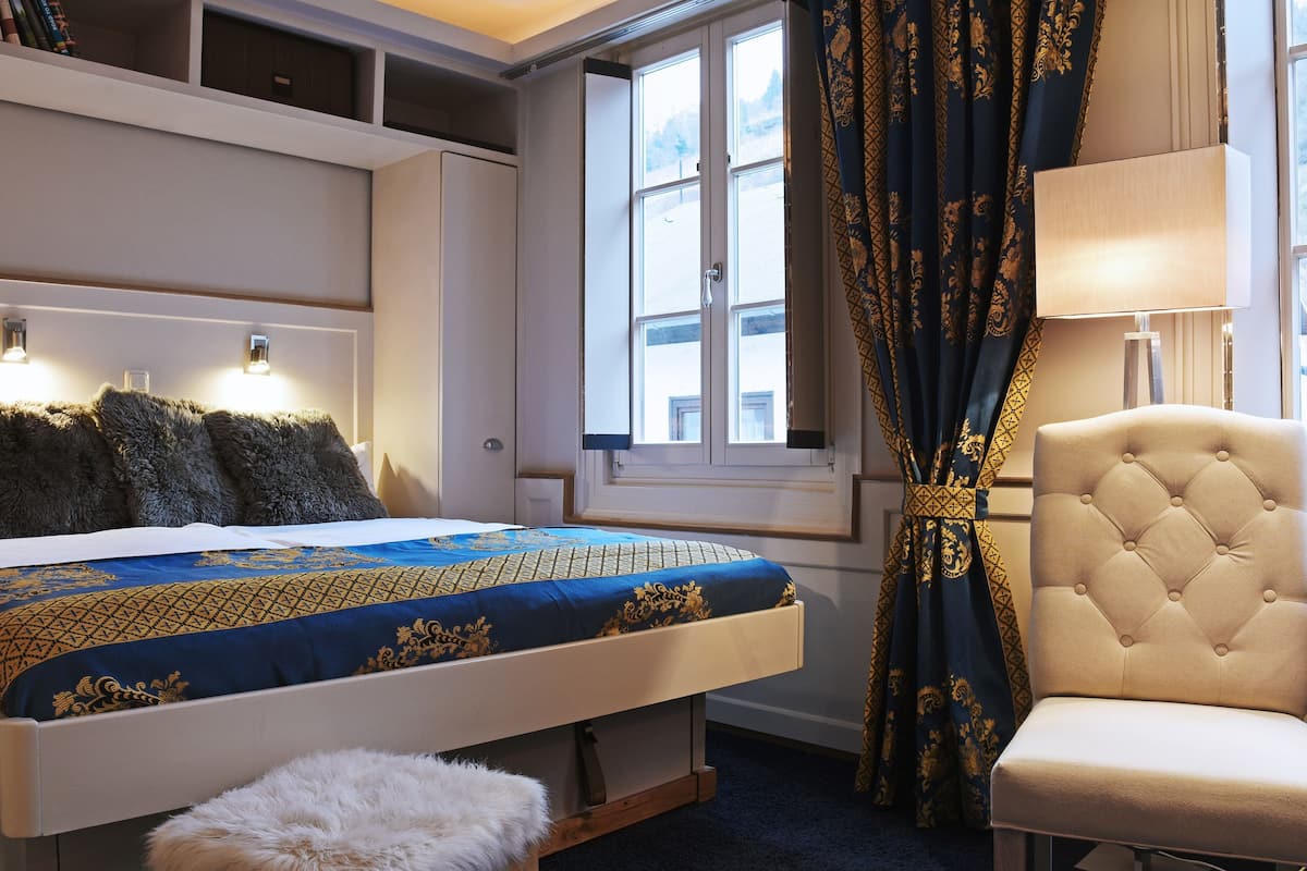 A bedroom with a bed, a chair, a window with curtains, and built-in shelves.