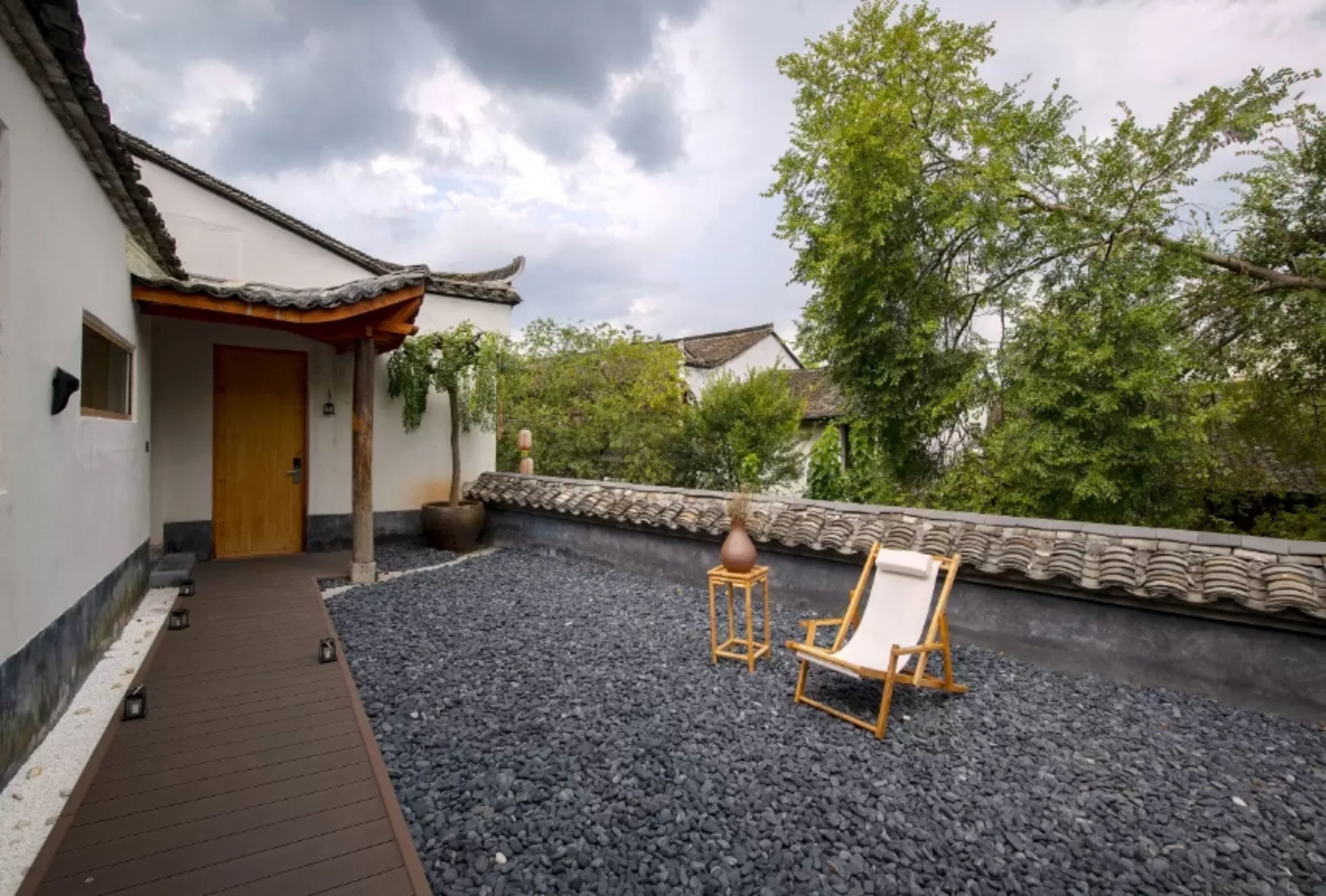 A traditional-style building with a tiled roof, a wooden door, and a small garden area with a gravel path, a chair, and a table.