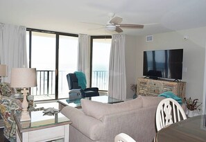 A living room with a sofa, a television, a ceiling fan, and a large window with curtains.