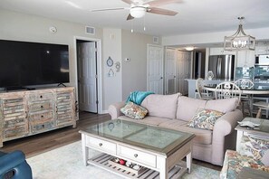 A living room with a television, a glass coffee table, a sofa, and a dining area with chairs and a chandelier.