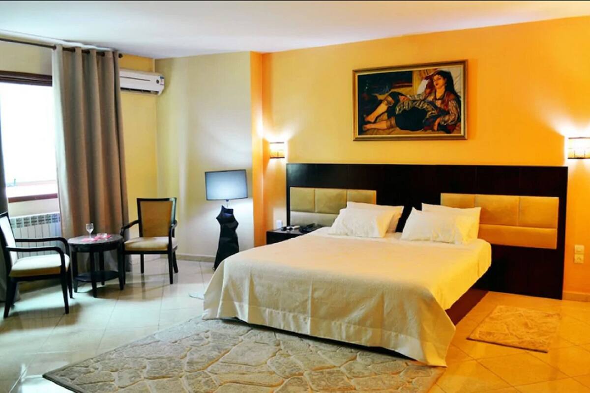 A hotel room with a bed, a chair, a table, and a painting on the wall.