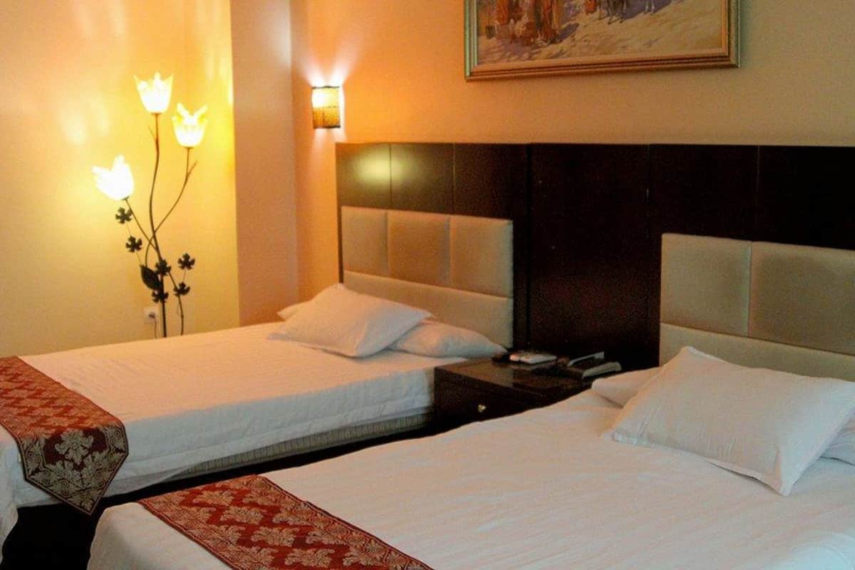 A hotel room with two beds, each with white linens and a headboard. There is a wall-mounted light fixture with a floral design and a painting above the beds.
