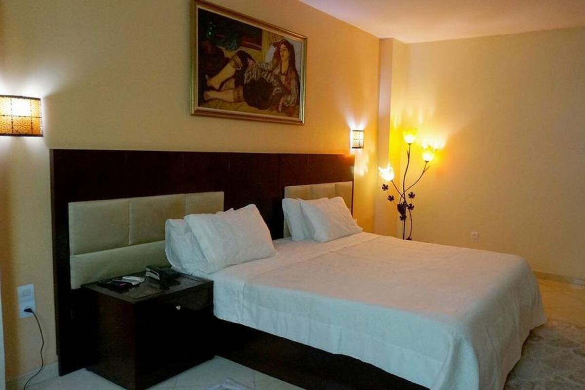 A hotel room with a large bed, a nightstand, a wall-mounted painting, and a wall-mounted light fixture.