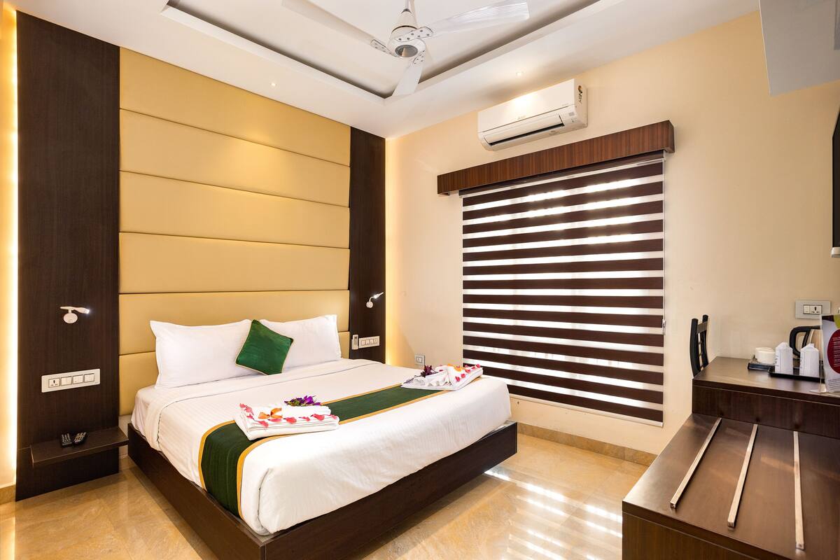 A hotel room with a large bed, a wooden headboard, a wall-mounted air conditioner, and a window with blinds.