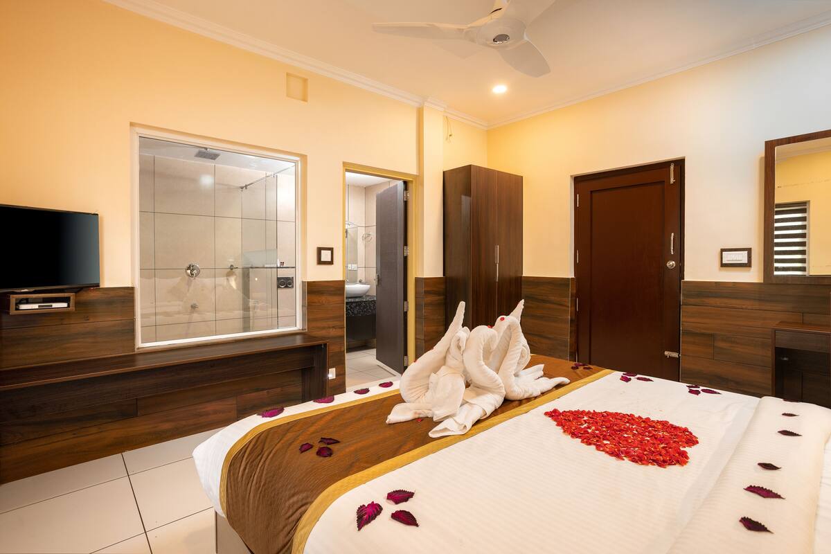 A hotel room with a large bed, a TV, a shower area, and wooden flooring.