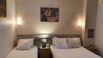 A hotel room with two single beds, a nightstand with a lamp, and artwork on the walls.