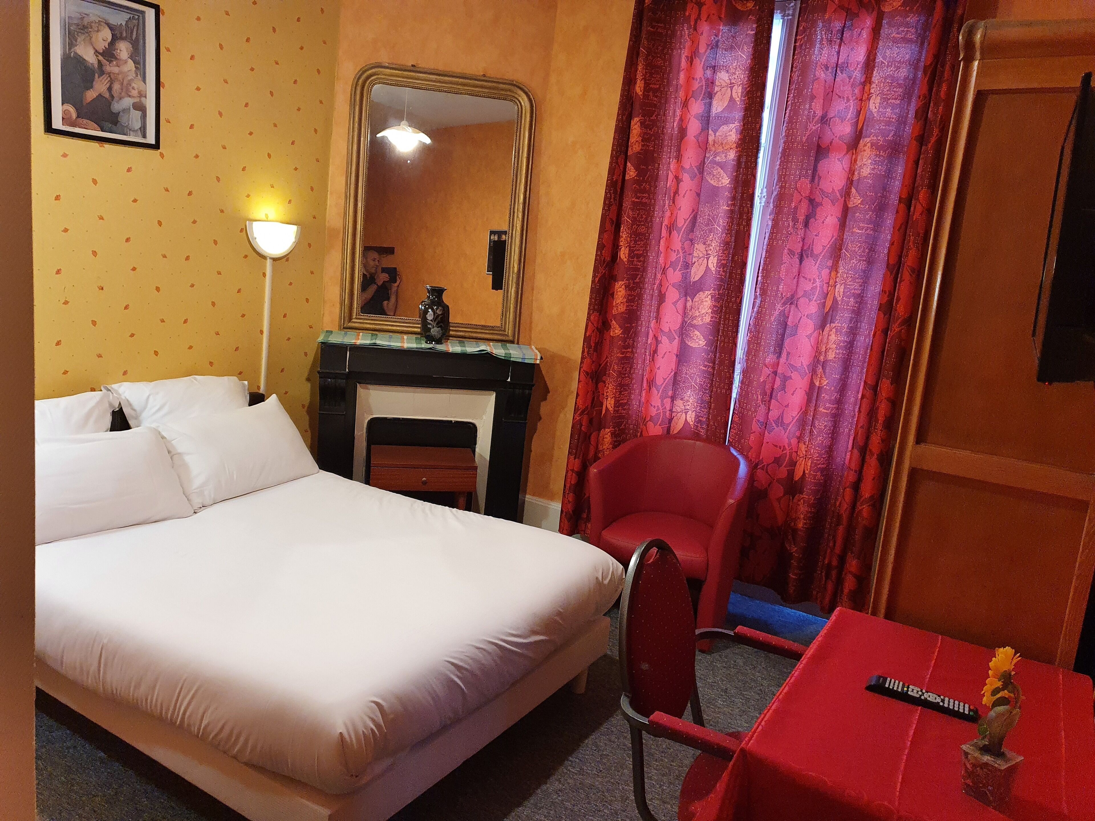 A hotel room with a bed, a red armchair, a small table, a mirror, and a fireplace.