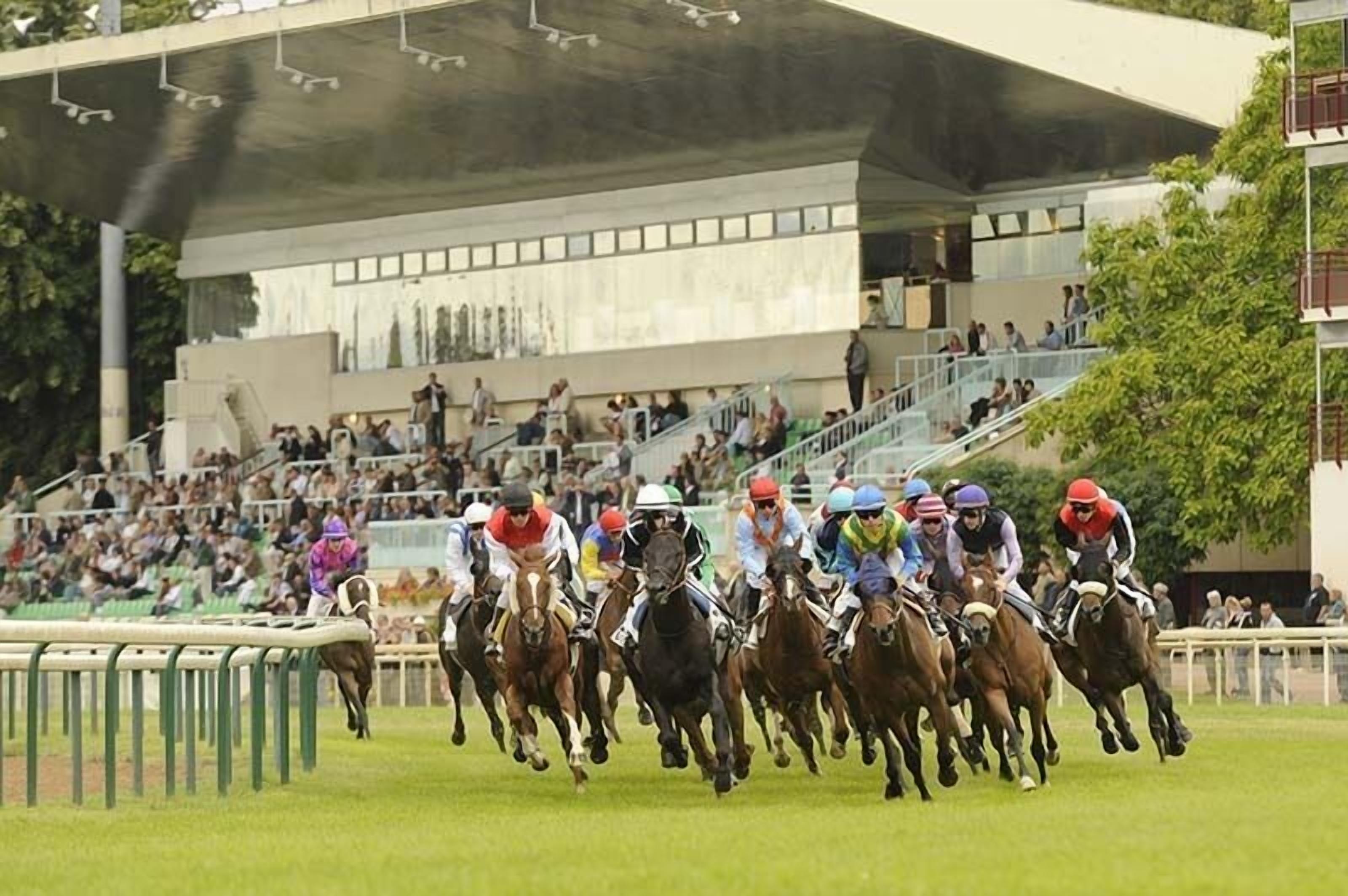 Carrera de caballos con jinetes y ejemplares en movimiento, espectadores al fondo y una estructura de edificio moderno.