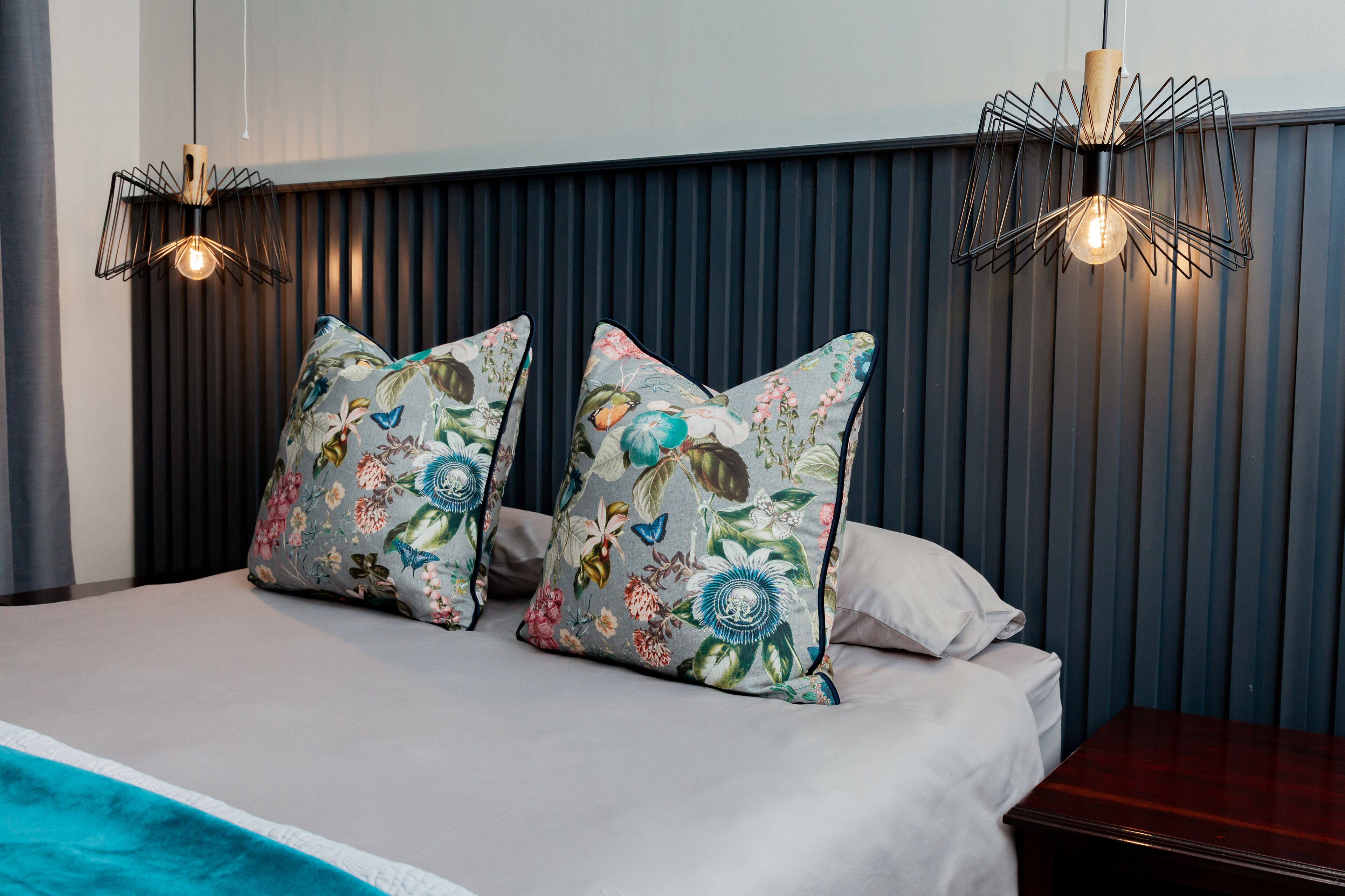 A neatly made bed with floral pillows, a wooden nightstand, and two pendant lights.
