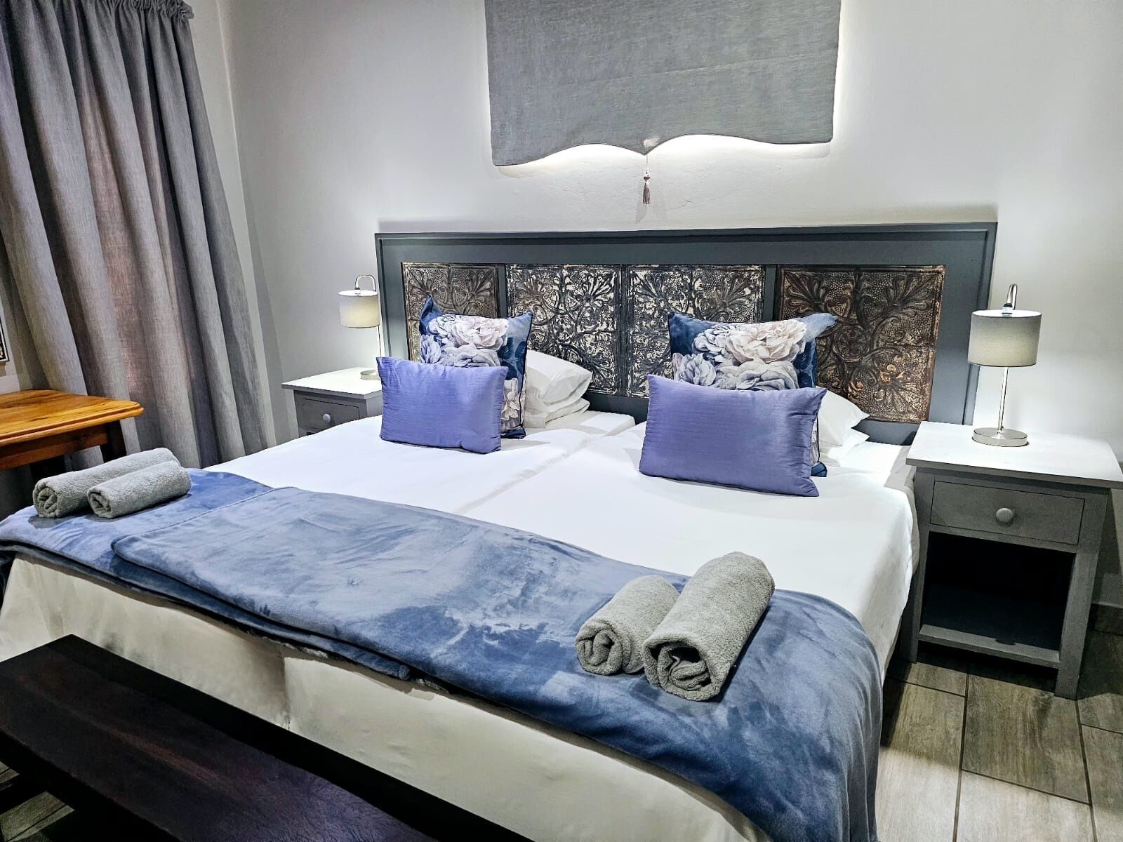 Cama bem arrumada com edredom azul e branco, duas almofadas e toalhas dobradas. Um criado-mudo com luminária e gaveta. O quarto tem cabeceira moderna e uma grande janela com cortinas.