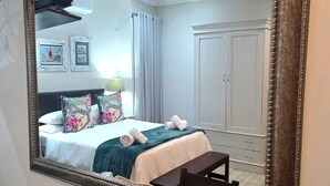 A hotel room with a large mirror, a bed with floral pillows, a bedside table with a tray of items, and a wardrobe. - 2 Owls Guesthouse (Potchefstroom)