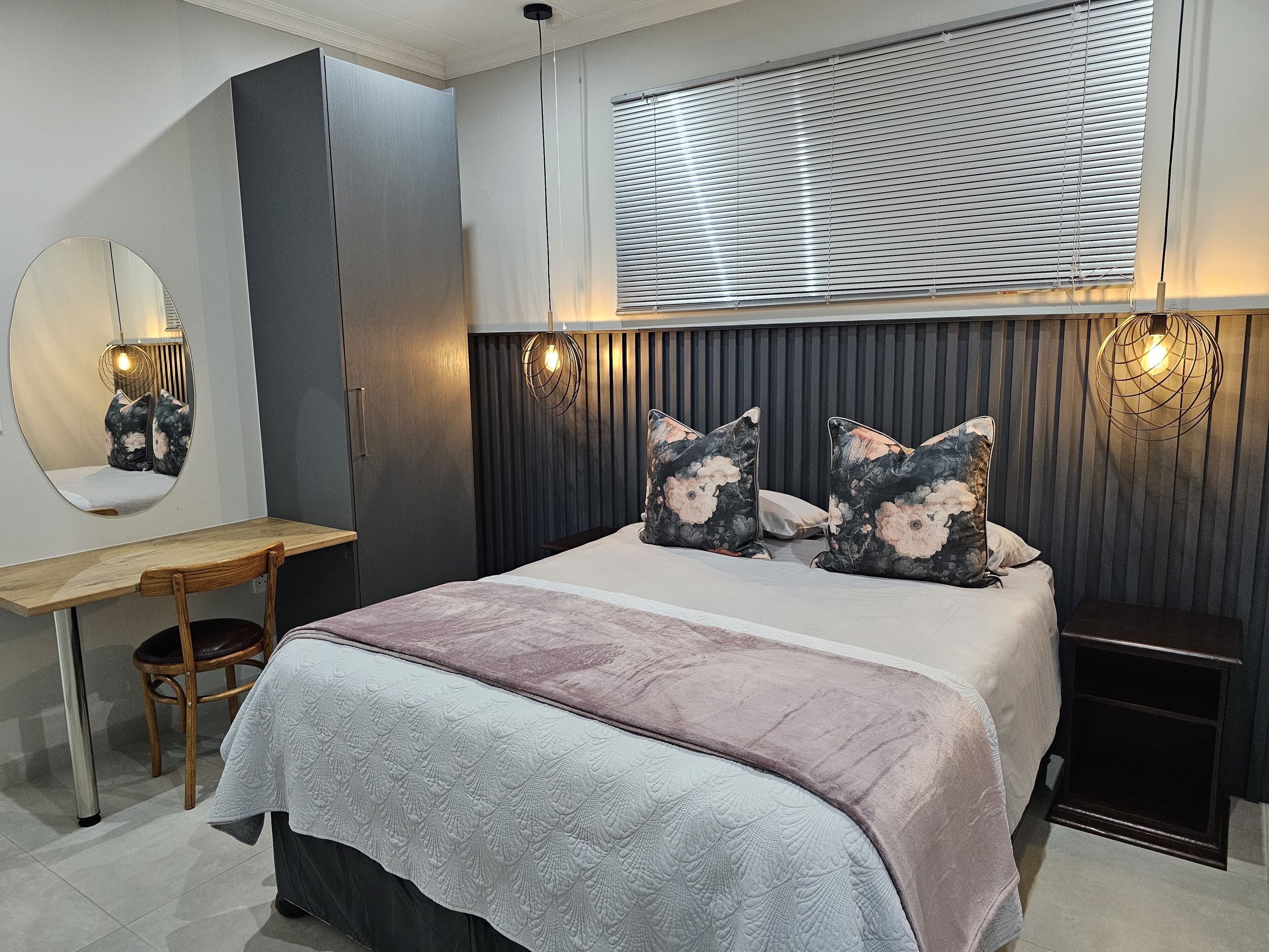 A bedroom with a bed, a desk, a chair, a mirror, and a window with blinds.