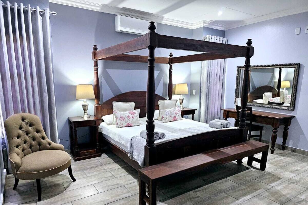 A four-poster bed with a wooden frame, a tufted armchair, a bedside table with a lamp, and a large mirror on the wall.