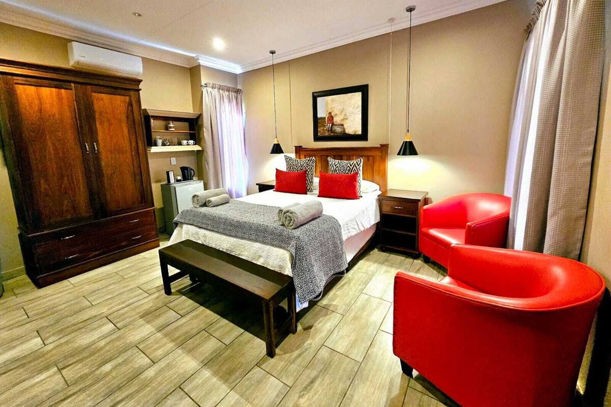A hotel room with a bed, a red armchair, a wooden wardrobe, a desk, and a painting on the wall.