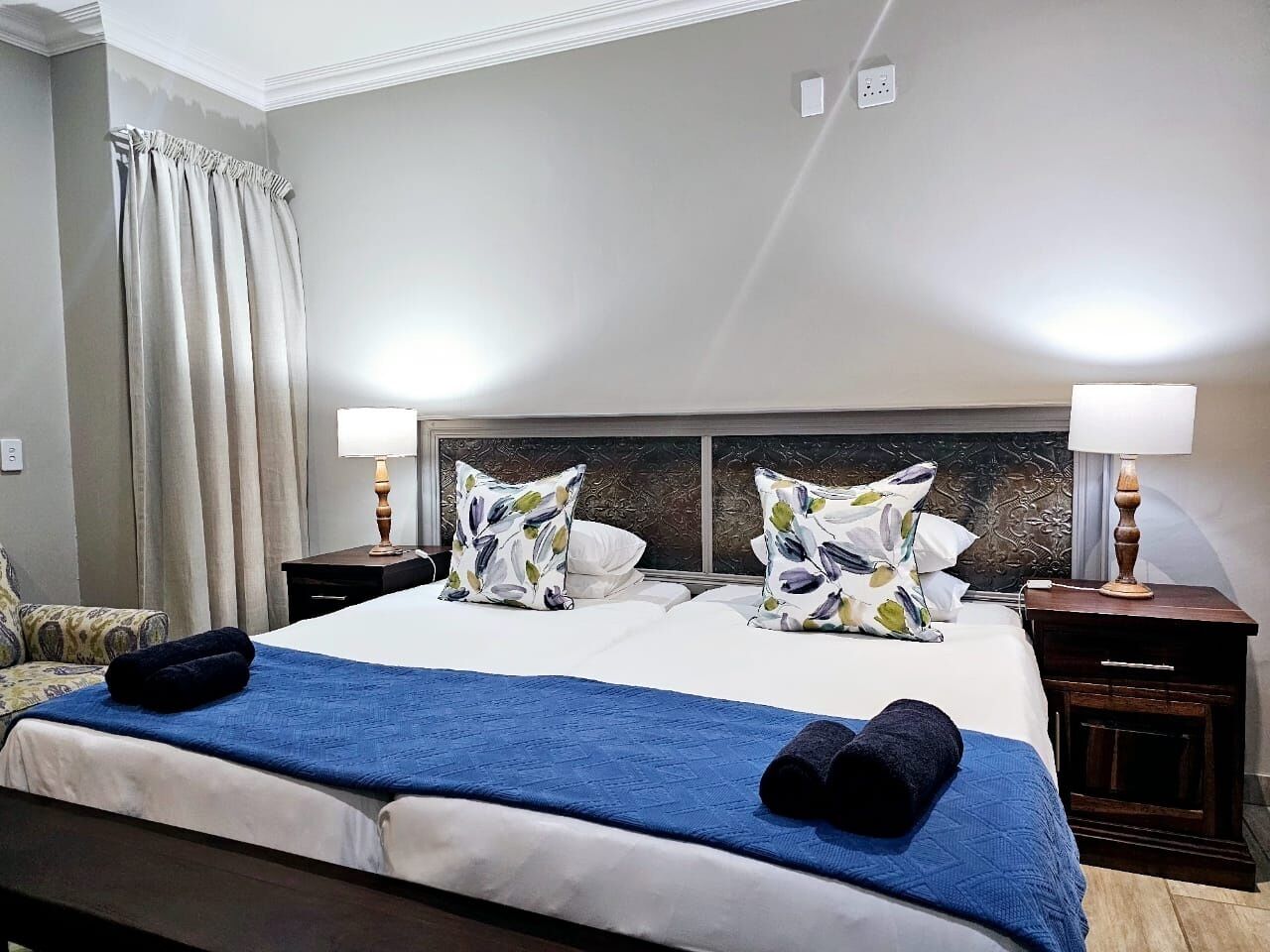 Une chambre d’hôtel avec un grand lit, deux tables de chevet avec des lampes, une tête de lit ornée d’un panneau décoratif et une fenêtre avec des rideaux.