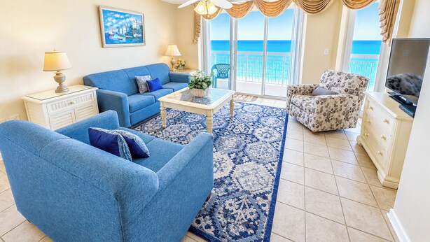 Ein Wohnzimmer mit einem blauen Sofa, einem gemusterten Teppich, einem Deckenventilator und Meerblick durch große Fenster.