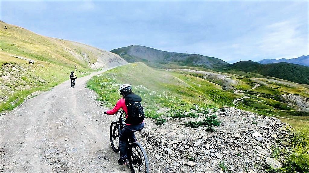 Un chemin de gravier où l’on aperçoit des cyclistes, entouré de collines verdoyantes et de montagnes.
