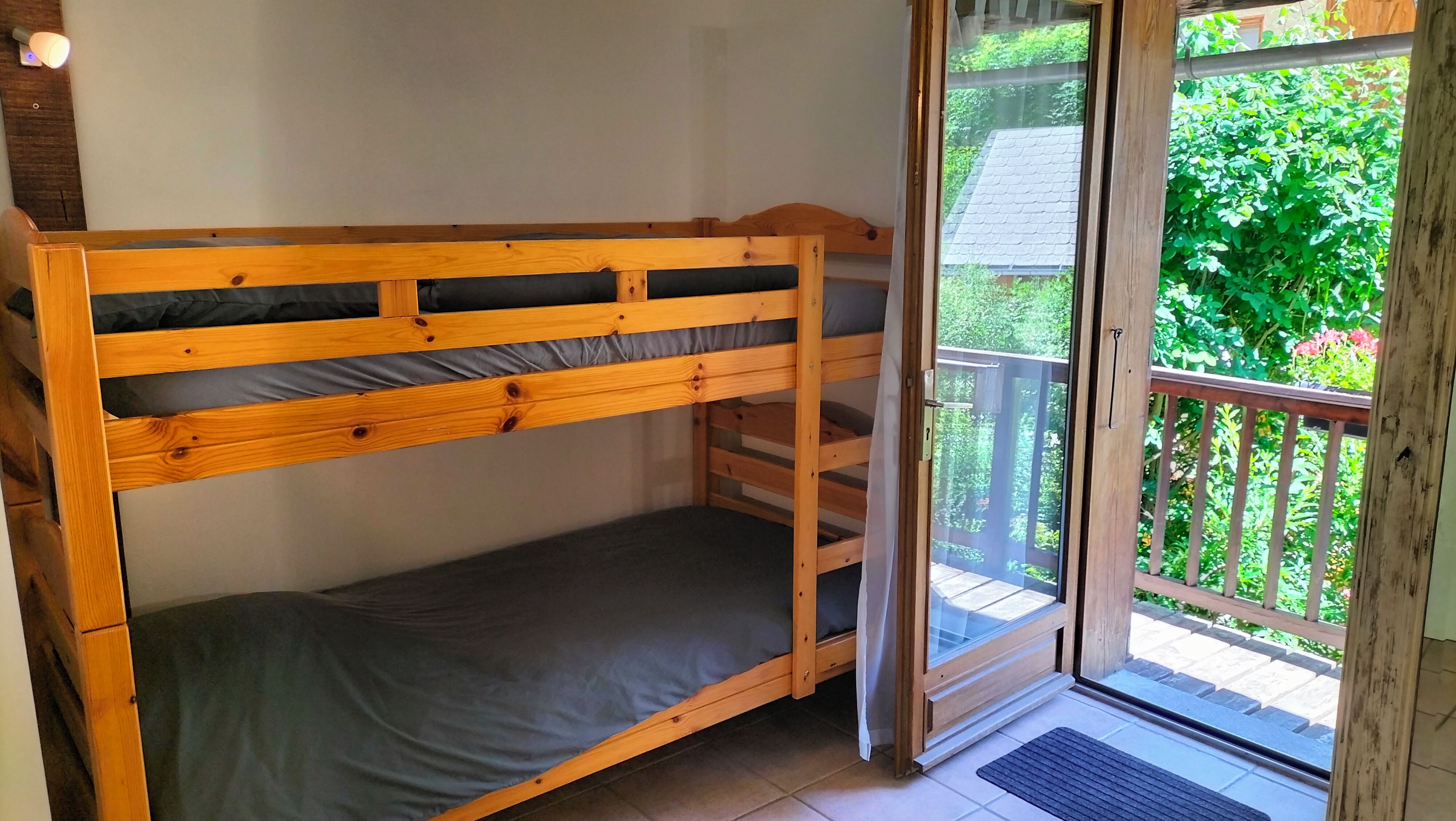 Una cama litera con estructura de madera y ropa de cama gris, una puerta corrediza de vidrio que da a un área exterior con terraza de madera y follaje.