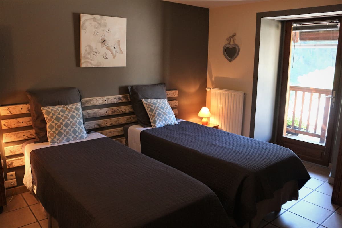 A room with two beds, a wooden headboard, a wall-mounted artwork, a window with a view, and a door leading outside.