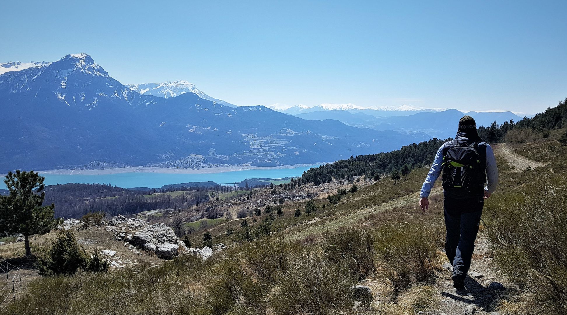 A hiker on a trail with a mountainous backdrop and a lake.