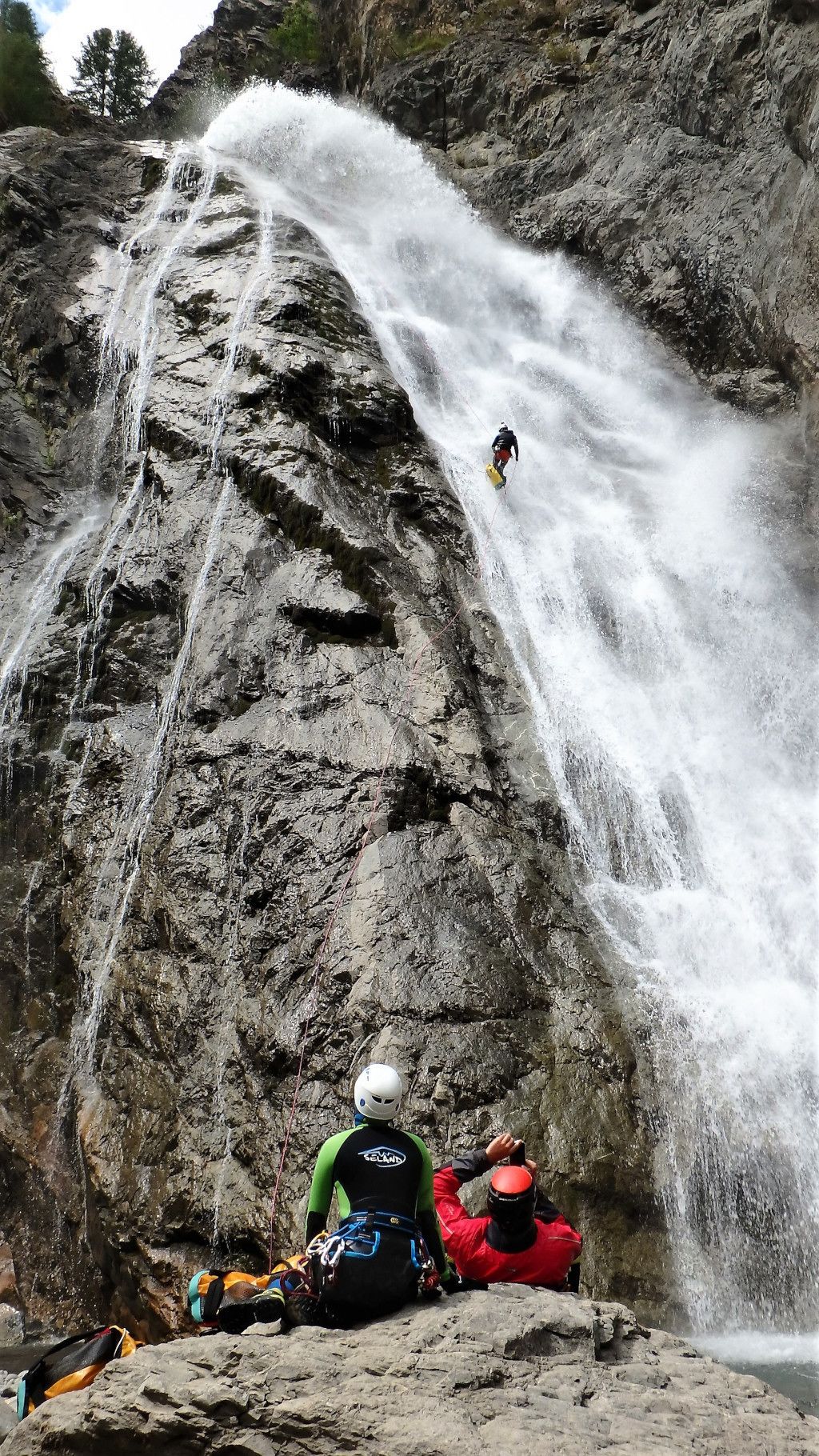 Una persona descendiendo en rappel por una cascada, con un grupo de personas sentadas abajo.