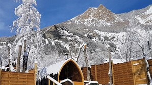 Ein mit Schnee bedecktes Holzhaus mit einem Steinkamin, umgeben von schneebedeckten Bäumen und Bergen im Hintergrund.