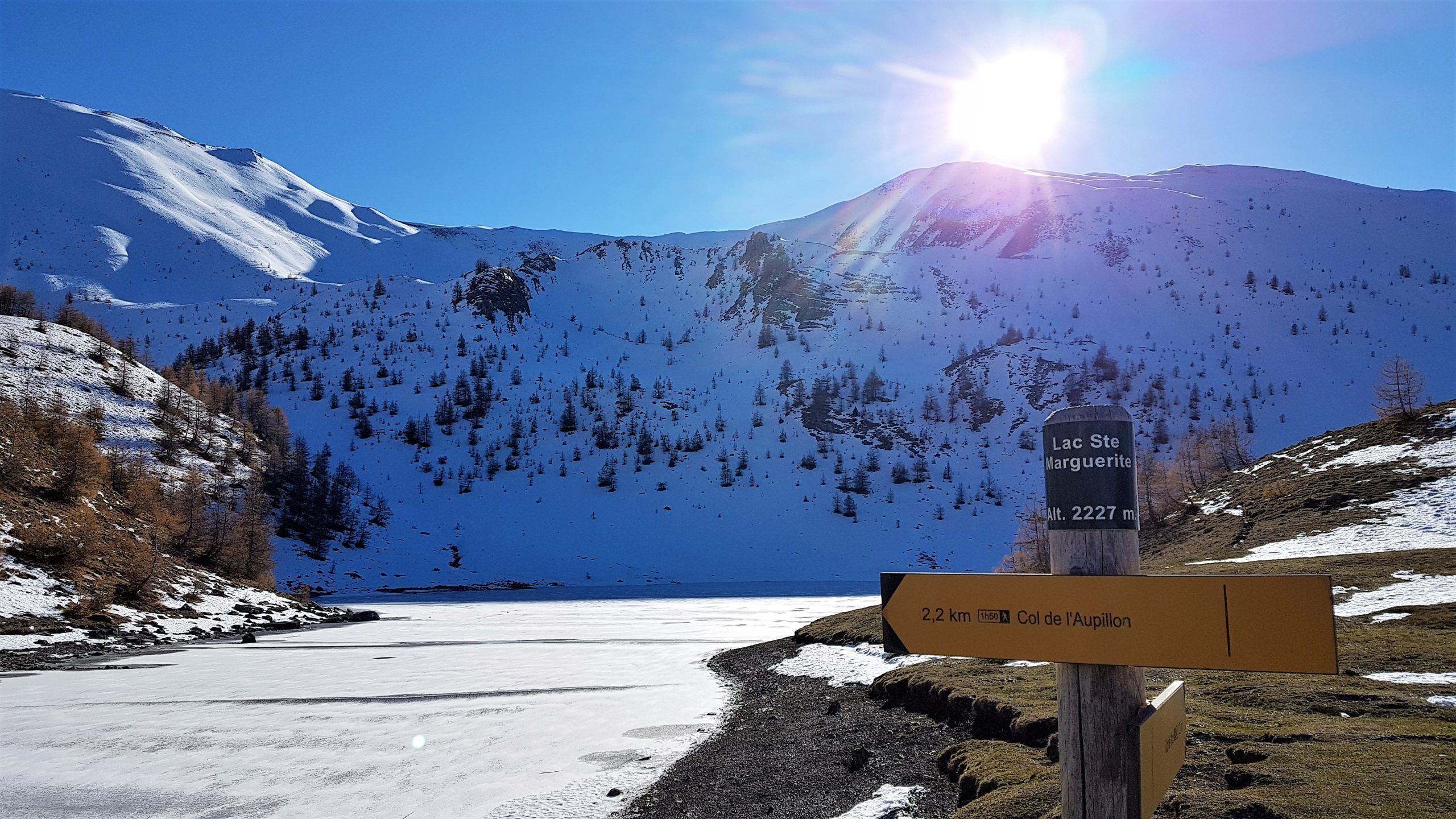 A snowy mountain landscape with a clear sky. There are signs indicating 'Lac Ste Marguerite' and 'Col de l'Aupillon'.
