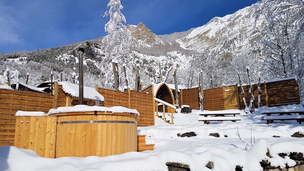 Ein hölzerner Wellnessbereich mit Whirlpool und Steinwänden, umgeben von schneebedeckten Bäumen und Bergen.