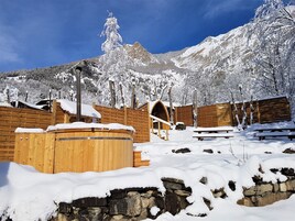 Un espace de détente en bois, doté d’un bain à remous et de murs en pierre, entouré d’arbres et de montagnes enneigés.