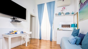 A hotel room with a flat-screen TV mounted on the wall, a dining table with breakfast items, a sofa with blue cushions, and a window with blue curtains.