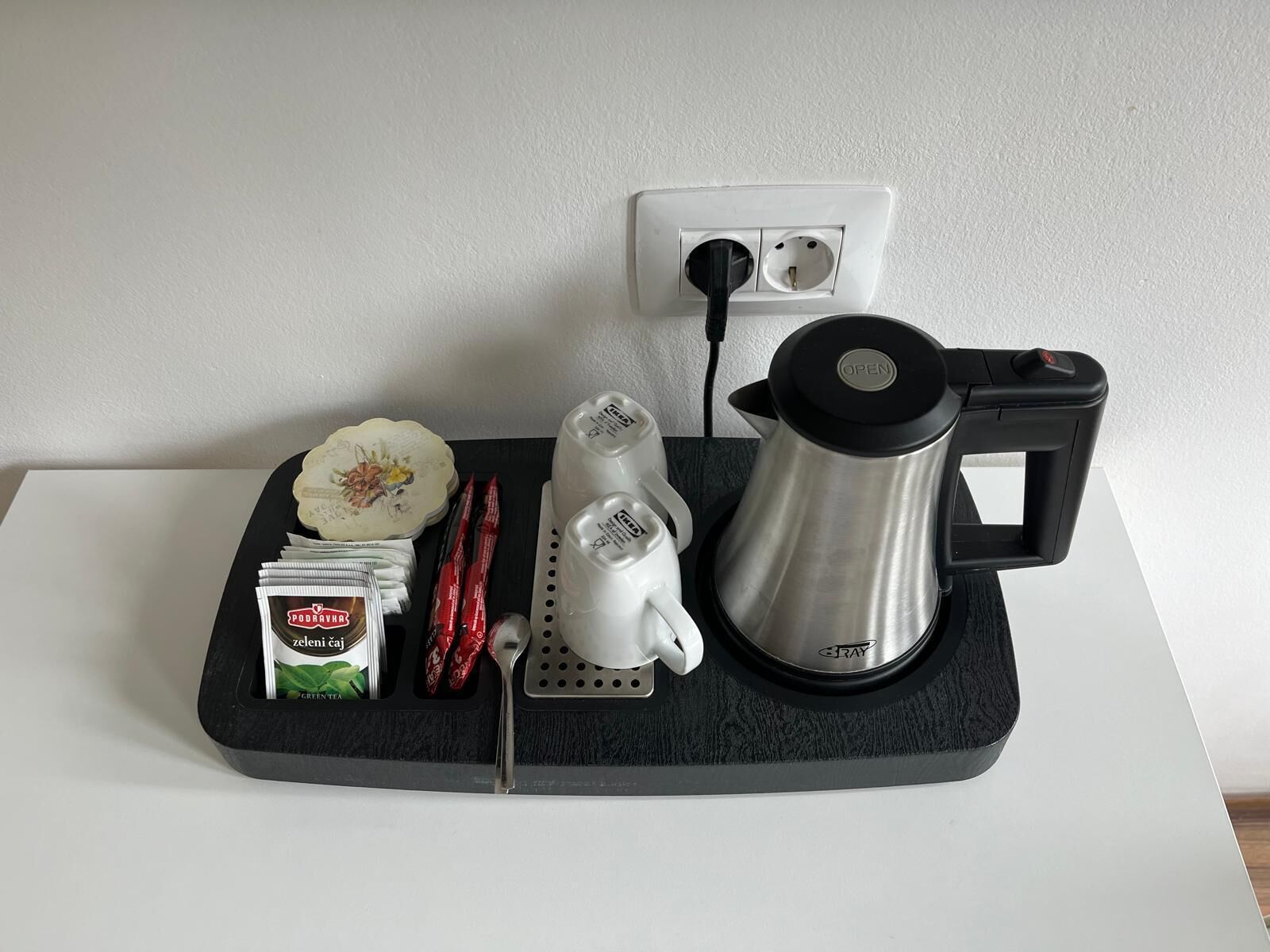 A kitchen setup with a coffee maker, a kettle, a spoon, a packet of tea, and a container of sugar on a black mat.