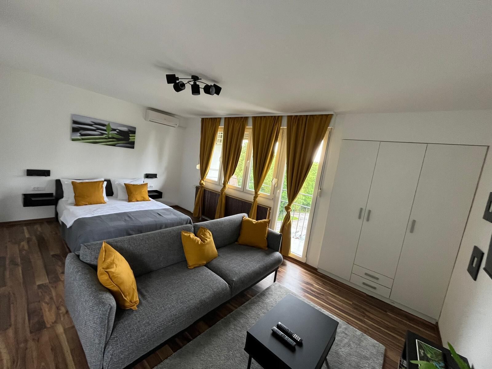 A modern bedroom with a bed, a grey sofa with yellow cushions, a small table with a remote control, and a window with curtains.