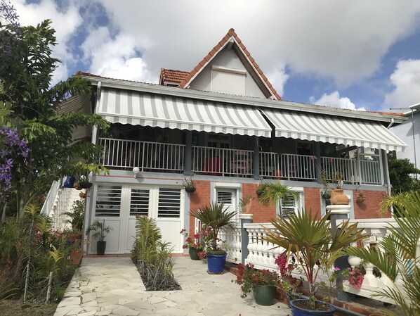 A two-story building with a balcony, potted plants, and a stone pathway. - Studio in villa with pool, garden and vehicle access at LAMENTIN in Martinique (Lamentin)