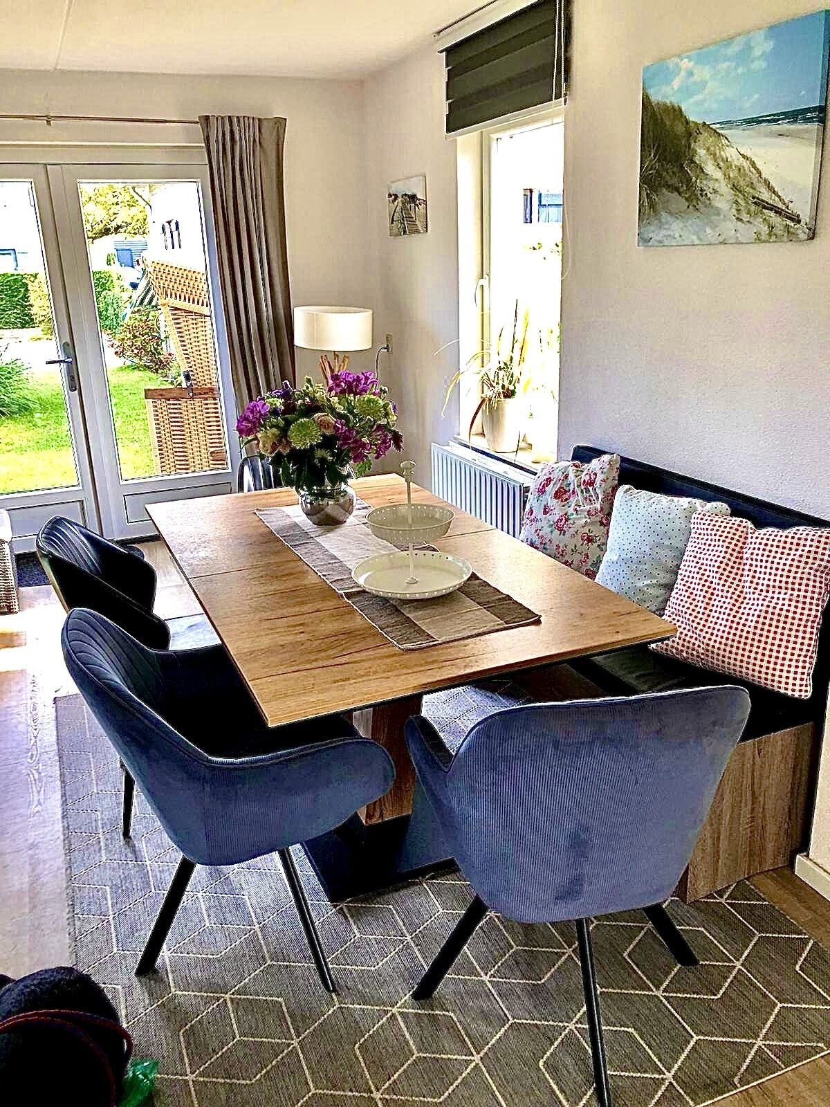 Een eetgedeelte met een houten tafel, twee blauwe stoelen, een bloemenvaas en een schilderij aan de muur.