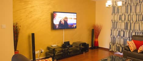A living room with a flat-screen TV mounted on the wall, a glass coffee table, a sofa with patterned cushions, and a patterned curtain.