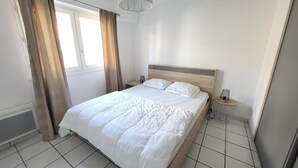 A hotel room with a bed, a nightstand, a lamp, and a window with curtains. - Cozy 1BR Apartment with Terrace, 200m from Sandy Beach (Les Sables-d'Olonne)