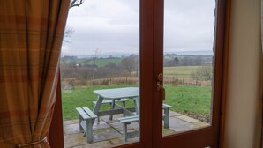 A view through a glass door of a picnic table set on a stone patio, with a rural landscape in the background. - Tanglewood Barn (Carlisle)