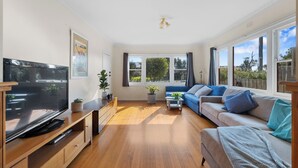 A modern living room with a flat-screen TV, wooden furniture, a blue sofa, and large windows offering a view of greenery. - Settle In Retro cottage, Cowes family holiday 10 min walk to beach, FREE WIFI (Cowes)