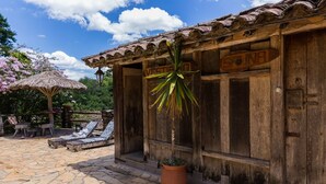 Un bâtiment rustique en bois, doté d’un toit de chaume, une terrasse en pierre avec des chaises longues et un pot de fleurs devant l’entrée.