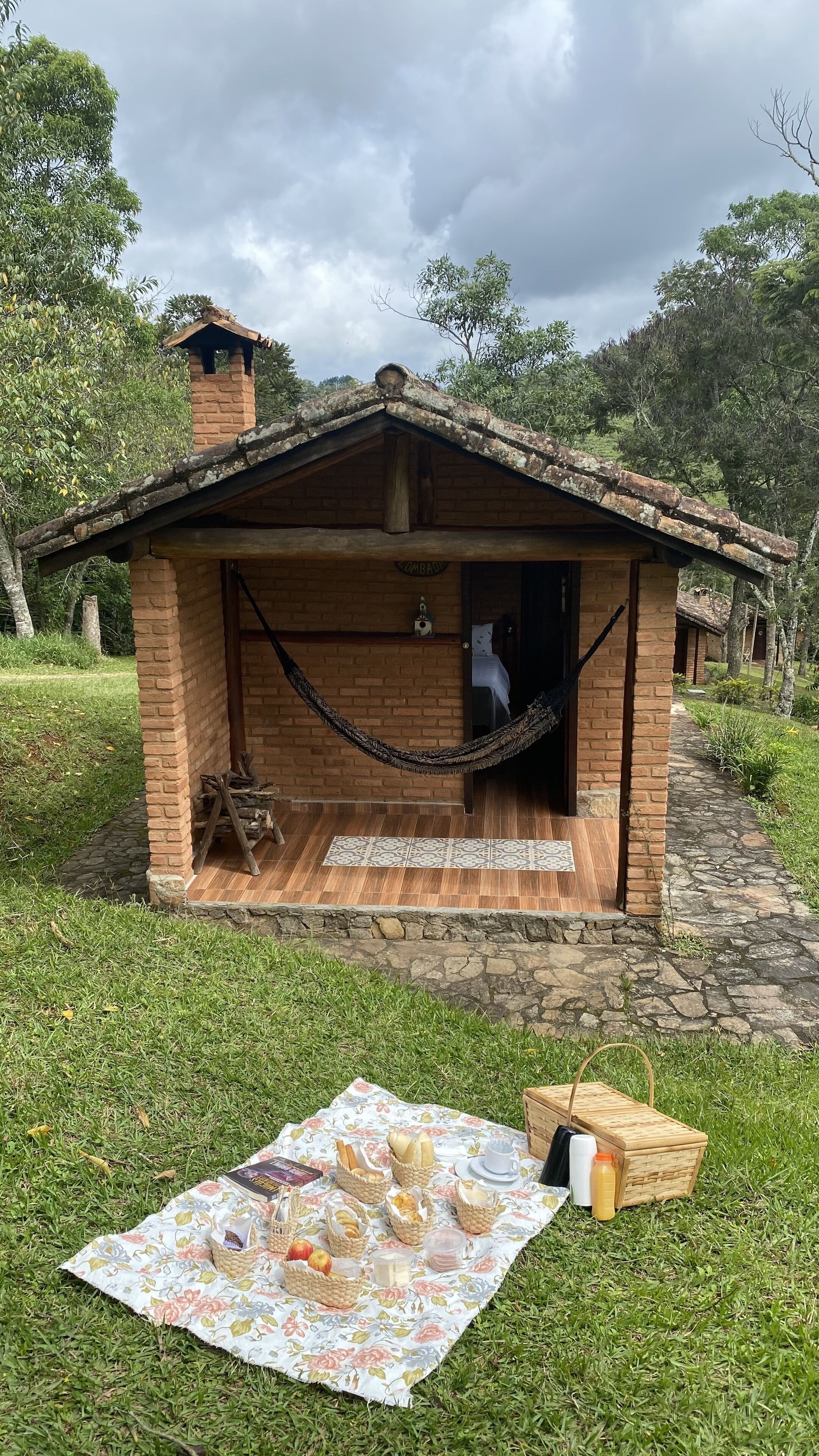 A rustic wooden cabin with a brick chimney, a hammock, and a picnic setup on the grass.
