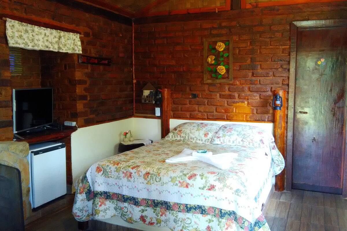 Quarto com parede de tijolos, cama com roupa de cama floral, televisão sobre suporte e porta com decoração floral.