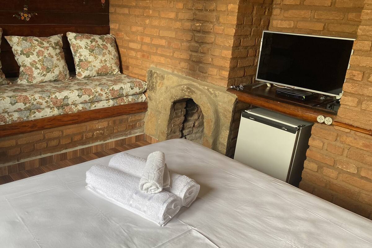 Quarto de hotel com cama, sofá com almofadas florais, televisão e pequena geladeira.